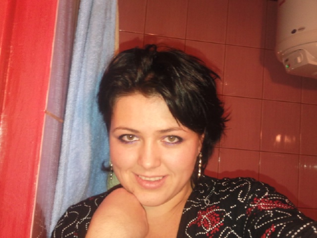 Uśmiechnięta kobieta z krótkimi, ciemnymi włosami i kolczykami, pozująca na tle łazienki z czerwonymi płytkami i zasłoną prysznicową.